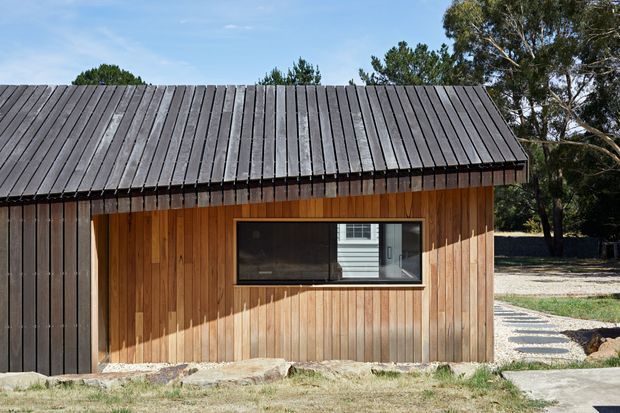Exterior view of a compact single-storey building with a steep pitched roof and vertically clad timber walls. The dark roof and matching dark vertical end cladding contrast with warm natural timber wall boards. A horizontal window is set into the facade, with a gravel path and landscaped surroundings visible to the side.
