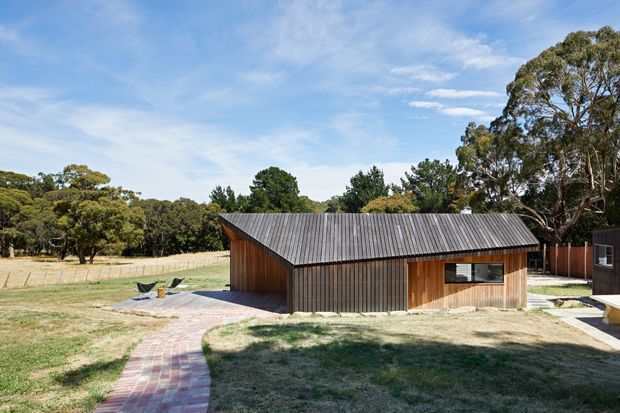 A single-storey rural outbuilding with a simple gabled form, vertical timber cladding, and a corrugated metal pitched roof. The building sits in an open landscaped setting with mature trees, dry grass, a brick-paved path, and a small paved outdoor area with seating. The composition has a clean, contemporary-rustic character under a bright blue sky.