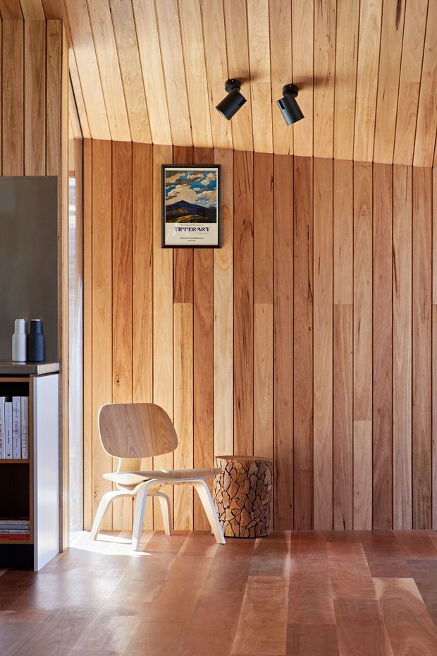 A warm, compact interior corner with extensive vertical timber panelling across the wall and sloped ceiling, complemented by a timber floor. A sculptural light-wood lounge chair sits beside a small round timber side table or stump, with a black built-in shelving unit and books at left. Black adjustable spotlights illuminate the wall, which also displays a framed poster. Natural light enters from a narrow window or glazed opening.