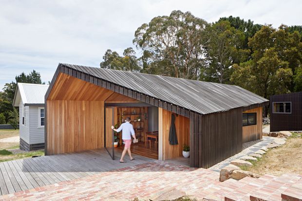 Exterior view of a compact contemporary timber-clad building with a steep pitched metal roof, large open glazed entry, and adjoining small structures. A timber deck and recycled-looking brick-paved path lead to the entrance, with native trees surrounding the site.