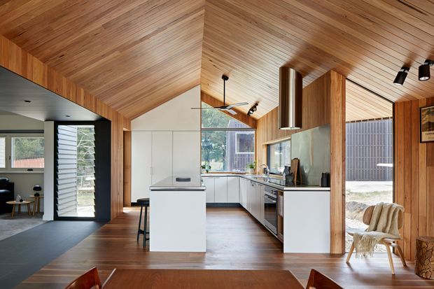 A contemporary open-plan kitchen and dining area with extensive warm timber lining across the pitched ceiling and walls. The kitchen features white flat-panel cabinetry, a central island, stainless-steel appliances and a dark benchtop, with large windows and glazed doors connecting to the outdoors. A dining table occupies the foreground, while a sitting area is visible through the open doorway on the left. The space has strong indoor-outdoor connection, clean joinery, and abundant natural light.