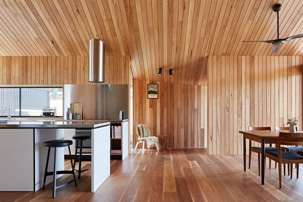 Open-plan interior with a compact kitchen and island breakfast bar opening into a dining and living area. The space is extensively lined in warm natural timber, including the vaulted ceiling, walls, and flooring. A minimalist kitchen features dark cabinetry, a pale island, stainless-steel appliances, and a cylindrical rangehood. A timber dining table and chairs sit beside large glazed openings, while a ceiling fan and track lighting provide functional lighting and ventilation.
