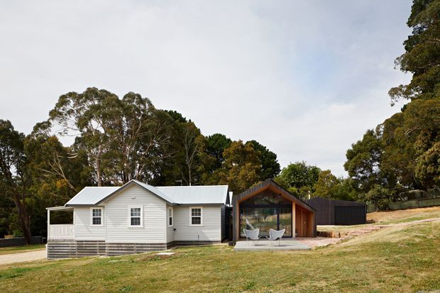 A low-rise residential property in a landscaped, wooded setting, combining a traditional white weatherboard cottage with a contemporary timber-framed glazed pavilion. Both structures have pitched metal roofs, with a dark-clad secondary outbuilding to the right. The scene is viewed from the exterior across a gently sloping lawn.