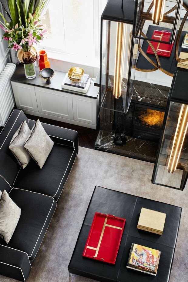 A sophisticated living room viewed from above, featuring a black sectional sofa with pale cushions, a large black upholstered ottoman coffee table, and a light neutral rug over dark timber flooring. A dramatic black marble fireplace with integrated reflective geometric shelving and warm vertical lighting forms the focal point. White panelled built-in cabinetry sits beneath a bright window, styled with flowers, books, and decorative objects. Red and gold accents add contrast to the monochromatic, luxurious scheme.