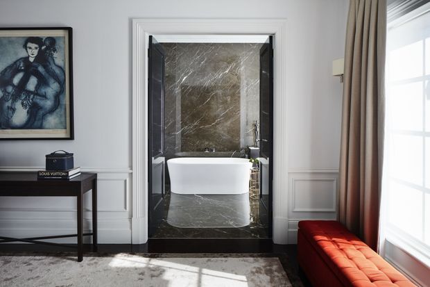 A refined bedroom or sitting area opens directly onto a luxurious bathroom through a wide doorway. The bathroom features a freestanding white bathtub, dark marble flooring and wall cladding, and black-framed detailing. The surrounding room has white paneled walls, a dark console table, framed artwork, full-height beige curtains, and a vivid red upholstered bench by a large window.