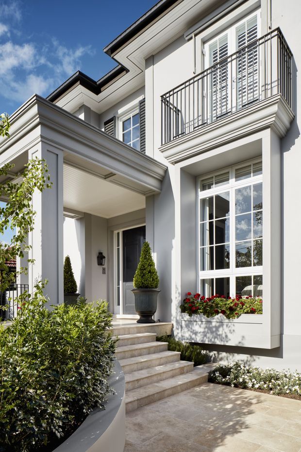 A bright exterior view of a refined multi-storey residence, focused on the formal front entry. The light rendered facade features strong classical detailing, projecting cornices, moulded trim, a covered porch with substantial square columns, a dark entry door, tall multi-pane windows with shutters, and an upper-level balcony with slender dark metal balustrades. Limestone or beige stone steps and paving lead through carefully landscaped planting with clipped topiary and flowering window boxes.