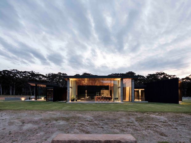 Wide exterior view of a low, contemporary single-storey residence set within a landscaped clearing and surrounded by mature trees. The home has a strongly horizontal composition, dark black cladding, extensive floor-to-ceiling glazing, and a central illuminated living area with a warm timber-lined ceiling and visible dining furniture. A covered outdoor area and lawn extend around the building, with an overcast evening sky creating a subdued atmosphere.
