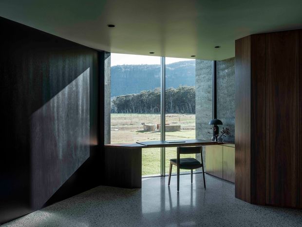 A dark, refined contemporary kitchen or compact living workspace with extensive floor-to-ceiling glazing overlooking a rural landscape and wooded hills. Deep timber wall panelling and tall timber cabinetry frame a muted green-grey ceiling and polished terrazzo-like floor. A slim island or peninsula with integrated sink, dark countertop, stool, and sculptural task lamp sits by the windows, creating a calm, highly composed interior with strong contrast between the shadowed timber surfaces and bright outdoor view.