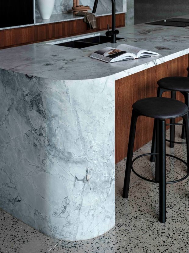 Close-up view of a contemporary kitchen island with a dramatically veined grey stone waterfall benchtop and rounded end. The island incorporates a dark undermount sink and matte black tapware, with warm timber cabinetry and black bar stools. A terrazzo floor and dark stone backsplash or wall surface complete the refined, material-focused composition.
