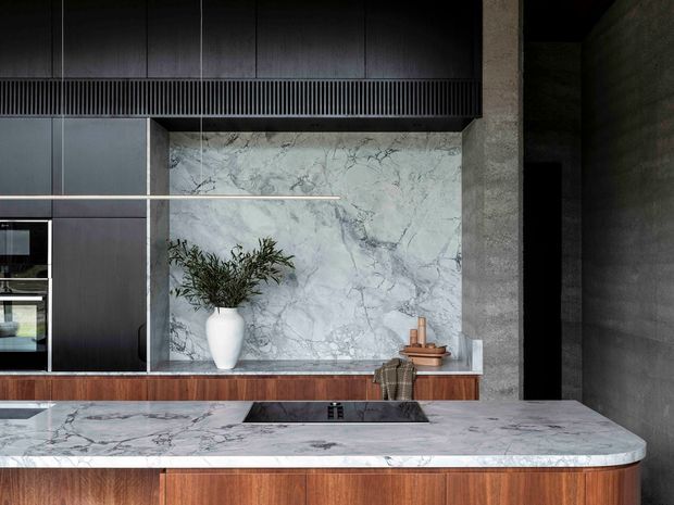 Close-up view of a sophisticated contemporary kitchen featuring dark vertical-panel cabinetry, integrated appliances, a large marble-look splashback, and a substantial island with a pale veined stone benchtop and rounded edge. Warm timber cabinetry below the splashback and island contrasts with dark textured wall surfaces, while understated decor and greenery add warmth.