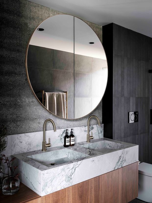 A sophisticated contemporary bathroom vanity featuring a large round frameless mirror, twin integrated basins, brushed brass gooseneck taps, and a dramatic white veined stone benchtop and splashback. The walls combine dark textured plaster with vertical dark timber or wood-look panelling, while the vanity cabinetry has a warm medium timber finish. A towel rail and wall-mounted flush plate are visible, creating a refined, moody interior.