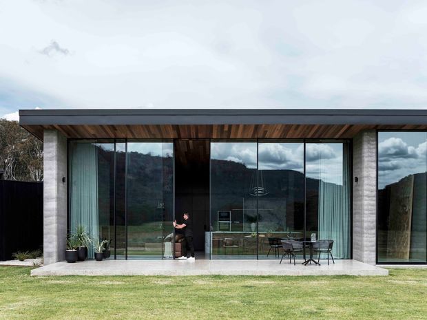Single-storey modern residence viewed from the lawn, with a broad flat roof, dark timber soffit, exposed concrete or pale masonry piers, and expansive floor-to-ceiling glazing. The glass reflects a wooded hillside and clouds while revealing a contemporary interior and a small outdoor dining setting on the terrace. The composition emphasises horizontal lines, transparency, and a strong connection between interior and landscape.