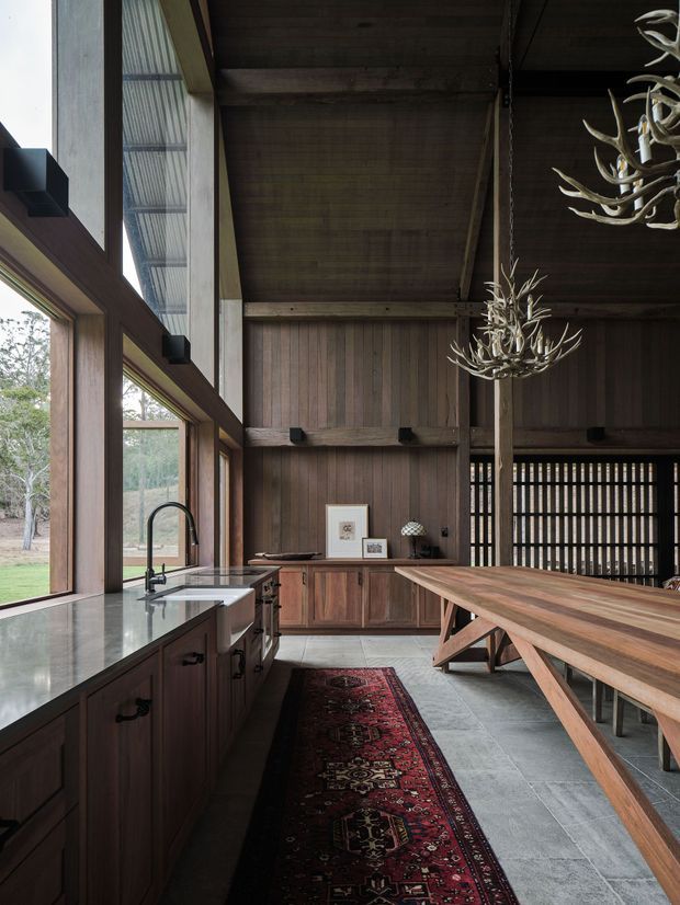 A dramatic, double-height kitchen and dining space with extensive floor-to-ceiling glazing, dark timber wall and ceiling lining, and exposed structural beams. The kitchen has dark timber cabinetry, pale stone benchtops, and black tapware. A long solid timber dining table with trestle-style supports occupies the right side, complemented by sculptural antler-like pendant lights and a patterned runner rug. The room opens to a leafy landscape through large windows and features a restrained, rustic contemporary material palette.