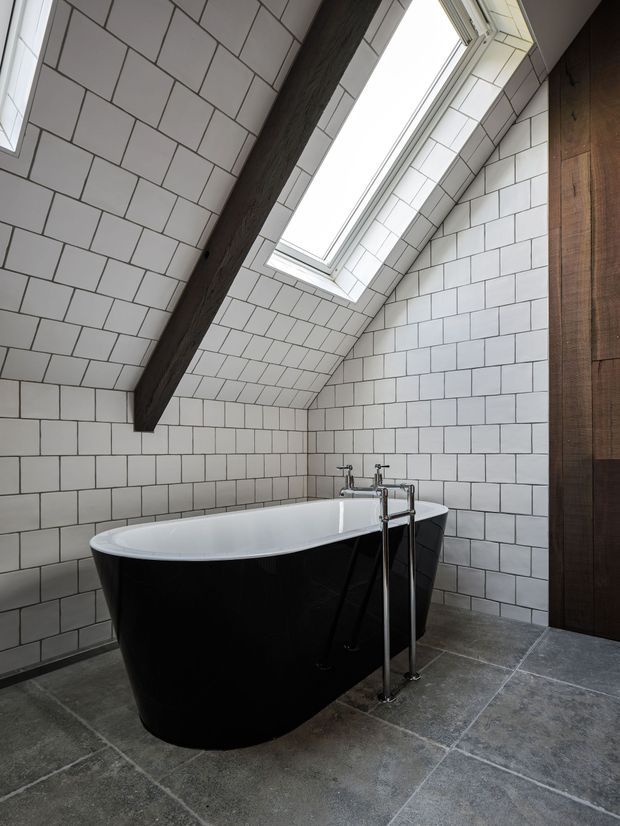 A dramatic attic bathroom with a freestanding black oval bathtub beneath a large skylight. White rectangular wall tiles cover the sloped walls and ceiling, contrasted by dark exposed timber beams and a dark timber door or panel. The floor is finished in large grey stone-look tiles, and the bath has a freestanding chrome tap and fixtures.