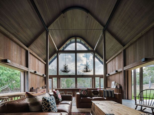 A rustic, lodge-like living room with a dramatic vaulted timber ceiling and exposed structural framing. Floor-to-ceiling glazing and a large arched window overlook dense greenery, while leather sofas, patterned cushions, a timber coffee table, and a rocking chair create a warm, relaxed interior.