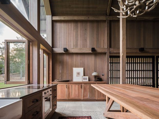 A spacious rustic kitchen with extensive timber wall panelling, exposed structural beams and a large timber dining island. Floor-to-ceiling glazing and an open window connect the room to a leafy outdoor setting. Dark timber cabinetry, integrated appliances, a stone benchtop and a sculptural branch-like pendant light create a warm, natural interior.