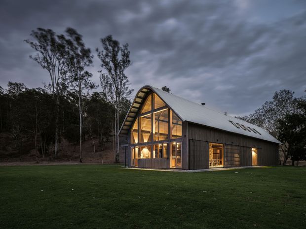 A dramatic dusk exterior photograph of a contemporary barn-like house set in a broad lawn and surrounded by native woodland. The building has a steep gabled roof with a curved or barrel-like gable profile, dark vertical timber cladding, extensive floor-to-roof glazing at the front, and warm interior lighting visible through the windows. Roof skylights and exposed structural framing are prominent.