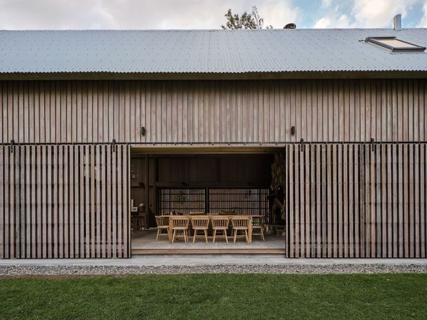 Front-on exterior view of a single-storey, barn-like building with a corrugated metal pitched roof and vertical timber cladding. Large timber slatted sliding doors are open, revealing a covered dining or entertaining area with a long timber table and chairs. A roof skylight and chimney are visible, with lawn and gravel in the foreground.