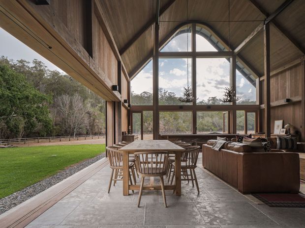 A spacious, timber-lined open-plan living and dining room with a dramatic vaulted gable ceiling and large arched-top windows. A dining table and chairs occupy the centre, while a substantial brown sectional sofa sits to the right. Full-height glazing opens the room toward a lawn and wooded landscape, creating a strong indoor-outdoor connection. The space uses exposed timber framing, dark timber window frames, and large-format grey stone or concrete-look floor tiles.
