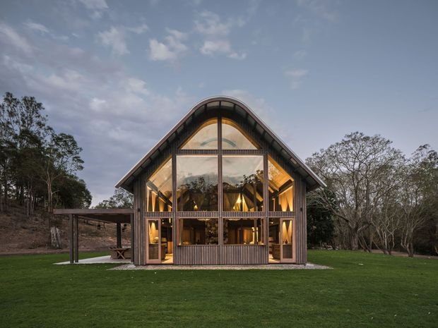 Front exterior view of a distinctive timber-clad, barn-like house set in a landscaped grassy clearing. The steep gabled form has a curved roof ridge, extensive floor-to-roof glazing, warm interior lighting, and a covered side structure. Mature trees surround the building, creating a rural setting at dusk.
