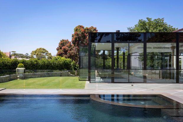 Exterior view of a contemporary glass pavilion beside a swimming pool. The pavilion has extensive black-framed floor-to-ceiling glazing, a flat roof, and a transparent glass balustrade overlooking a manicured lawn and landscaped suburban garden. A spa or shallow pool section is visible in the foreground.