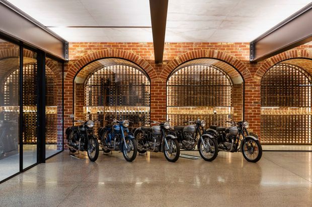 Interior commercial space with rows of motorcycles displayed in front of exposed red-brick arched walls and black metal security grilles. The polished concrete floor, dark-framed glass partitions, and exposed structural beam create an industrial showroom or adaptive-reuse character.