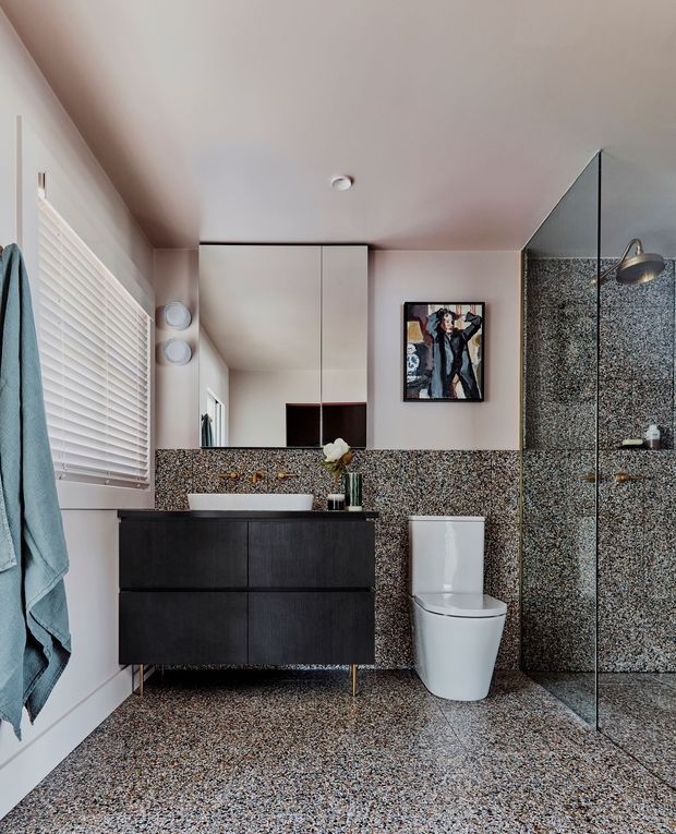 Contemporary bathroom with a wall-mounted dark timber vanity, white vessel basin, wall-mounted brass tapware, white toilet, and frameless glass shower enclosure. Speckled terrazzo-style flooring and matching shower wall finish create a bold continuous surface, complemented by white walls, a large mirrored cabinet, window blinds, and muted blue towels.