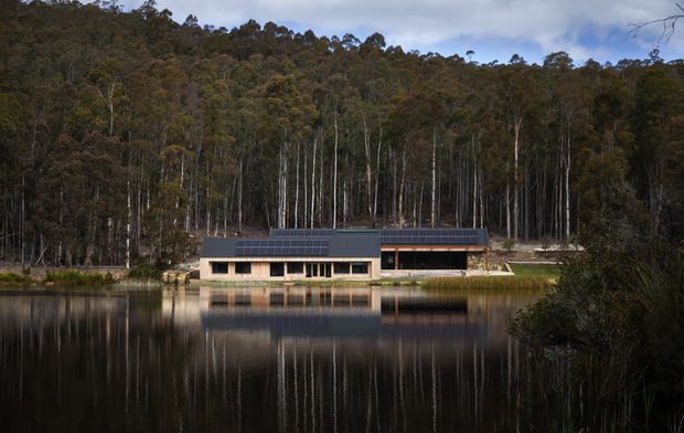 A long, low modern building sits beside a calm lake, with extensive dark roof-mounted solar panels and a forested hillside behind it. The facade combines pale masonry or rendered walls, dark window bands, and timber-toned cladding, all reflected in the water. The image is a moody architectural exterior photograph in a secluded natural setting.