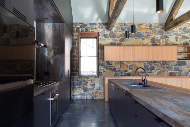 A contemporary rustic kitchen with dark integrated cabinetry, a large central island, timber wall-mounted storage, exposed rough-hewn ceiling beams, and a full-height irregular natural stone feature wall. Black pendant lights, stainless appliances, and a dark polished floor complete the high-contrast, earthy interior.