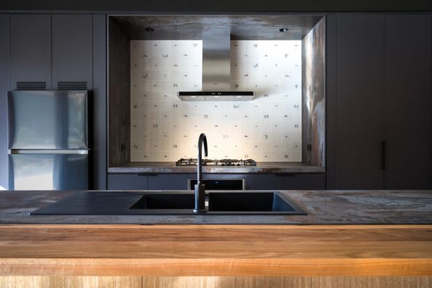 A dark, contemporary kitchen viewed across a large island. The foreground island has a warm timber-clad front and a dark stone-looking benchtop with an integrated sink and tall black tap. Behind it are dark matte cabinetry, a stainless-steel refrigerator, a cooktop, and a pale patterned tiled splashback framed by a darker stone or concrete recess. The mood is dramatic and refined, with warm timber contrasting against charcoal cabinetry.