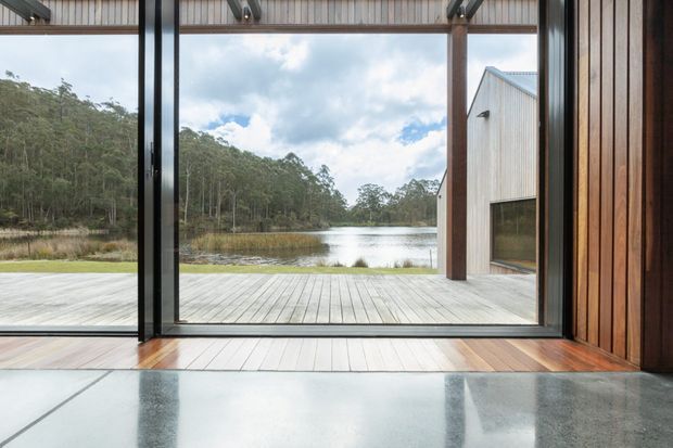 A refined interior view through expansive floor-to-ceiling glazing toward a timber deck, calm lake, and forested landscape. The room features polished concrete flooring, warm vertical timber wall cladding, exposed timber ceiling elements, and slim dark window and door frames. A simple pale timber-clad outbuilding is visible outside, reinforcing the contemporary natural setting.