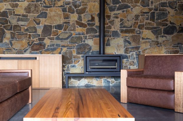 A cozy interior seating area centered on a contemporary black wood-burning fireplace set into an irregular natural stone feature wall. Two brown leather armchairs or sofas with pale timber frames flank a substantial warm timber coffee table, while low timber cabinetry is visible to the left. The room has a restrained, rustic-modern character with strong natural materials and dark flooring.
