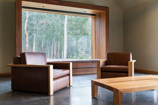 A refined sitting room with two substantial leather armchairs and a solid timber coffee table arranged around a built-in window seat. A large picture window frames a wooded landscape, while dark polished flooring, warm timber joinery, and neutral walls create a calm, nature-connected interior.