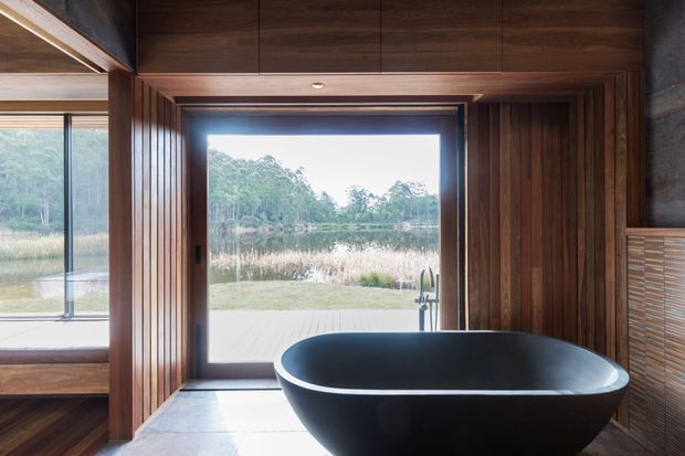 A refined bathroom interior featuring a large matte-black freestanding oval bathtub positioned in front of expansive glazing overlooking a tranquil lake and wooded landscape. Rich dark timber wall panelling and cabinetry surround the space, with a warm timber ceiling, recessed lighting, and a dark tiled floor. The room has a strong connection to nature and a dramatic yet serene material palette.