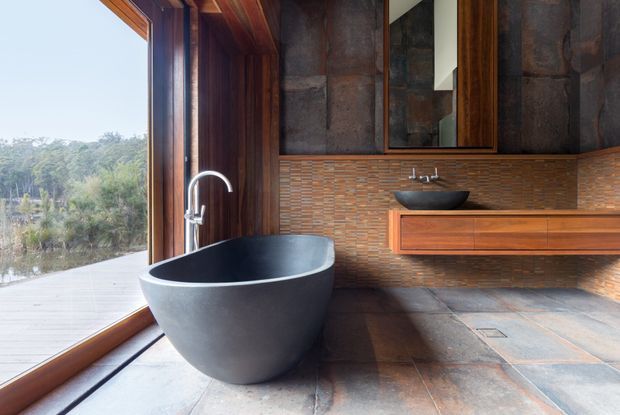 A warmly detailed contemporary bathroom with a freestanding dark stone bathtub beside full-height timber-framed glazing. The room features large slate-like floor and wall tiles, a textured horizontal mosaic tile backsplash, a floating timber vanity with a black vessel basin, and dark timber wall cabinetry. A tranquil leafy landscape is visible outside.
