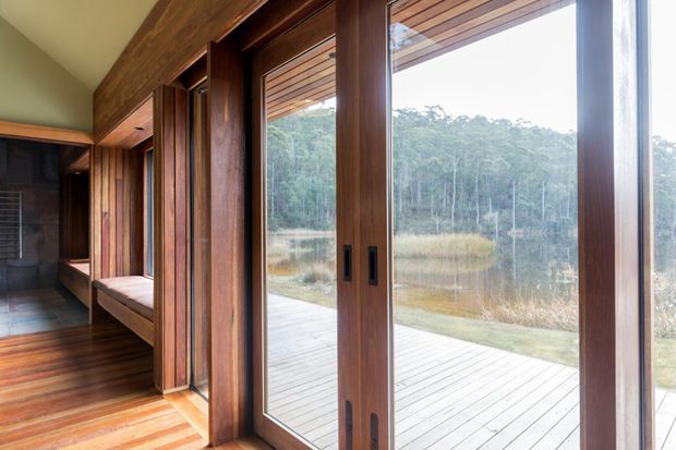 Interior view of a timber-lined room opening onto a broad outdoor deck through large floor-to-ceiling sliding glass doors. Rich dark timber framing, built-in window seating, timber flooring, and a covered timber soffit create a warm connection to the surrounding wetland and bush landscape.