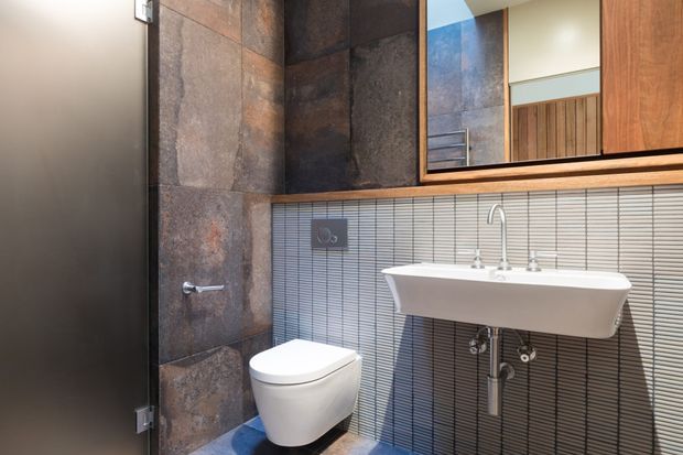 Compact contemporary bathroom featuring a wall-hung white toilet and a wall-mounted white basin with exposed chrome plumbing. The walls combine large rust-brown stone-look tiles with narrow horizontal grey tiles, while a large timber-framed mirror and timber shelf add warmth. A dark door is visible at left.