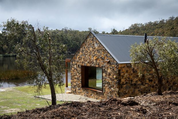A small stone-clad gable-roof building set beside a calm body of water and surrounded by dense bushland. The facade features irregular natural stonework, a dark metal pitched roof, and a large glazed opening on the front wall. The scene has a rugged, secluded bush setting under overcast light.