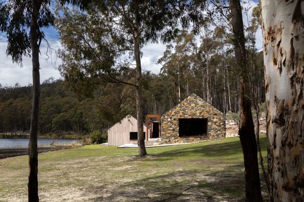 A low, contemporary pavilion-like building sits in a wooded lakeside setting. The main structure has a steep triangular gable facade clad in irregular multicoloured natural stone, with a large dark rectangular opening or glazed aperture. A smaller pale timber-clad gabled volume adjoins it, and a timber-framed entrance connects the forms. Eucalyptus trees, lawn, and the water beyond establish a secluded bush setting.
