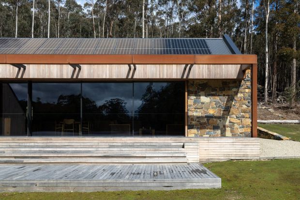 Single-storey contemporary pavilion-like building set among eucalyptus trees, viewed from the front across a broad timber deck. The low roof is covered with solar panels and edged with dark metal trim. Full-height dark glazed sliding doors span the main facade, with warm timber cladding and projecting angled sun-shading elements above. A prominent irregular natural-stone wall forms the right side, and wide timber steps lead down to the lawn.