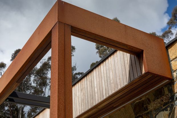 Low-angle architectural detail showing a bold rust-coloured rectangular steel portal frame projecting around a timber-clad building element. Dark steel framing, vertical weathered timber boards, and a rough stone wall are visible, with trees and a cloudy sky in the background. The composition emphasises strong geometric lines, exposed structural elements, and contrasting industrial and natural materials.