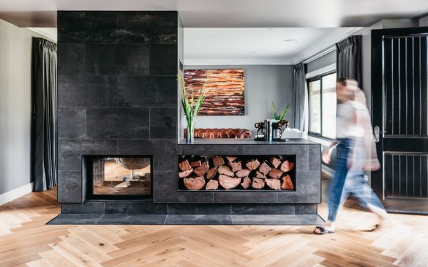 Contemporary living room centered on a large dark stone-clad fireplace with an integrated firebox and open firewood storage. The room has pale herringbone timber flooring, grey walls, full-height dark curtains, large glazing, a black-framed door, a leather sofa visible behind the fireplace, and a slim kitchen or bar counter. An abstract artwork and indoor plant add colour, while a person is captured in motion near the doorway.