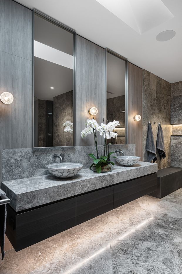 A luxurious contemporary bathroom featuring a long double vanity with dark wood cabinetry, a substantial grey natural-stone benchtop and matching vessel basins. Full-height mirrors are framed by vertically grained timber wall panels, while the walls and floor use richly veined stone. Warm accent lighting, orchids, and dark towels add refined detail. A skylight or recessed ceiling feature and a freestanding-style bath edge are visible in the reflection and at right.