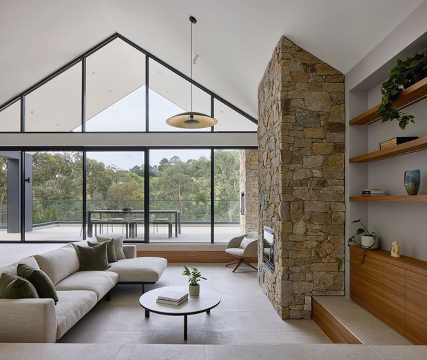 A bright, open-plan living room with a large sculptural stone fireplace wall, exposed pitched ceiling, and extensive black-framed floor-to-ceiling glazing opening to a balcony or terrace. The room features a pale sectional sofa, round coffee table, timber built-in shelving and cabinetry, and a simple pendant light. Views of trees are visible beyond the glazing.