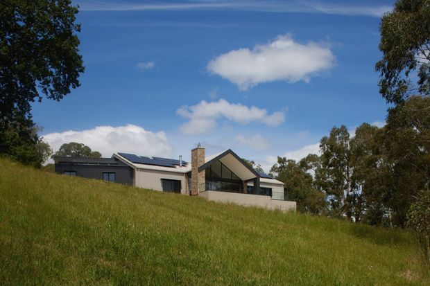 A contemporary single-storey house set on a gently sloping grassy site, viewed from a distance. The building has dark metal-clad volumes, a prominent pitched gable roof with a glazed front facade, a stone-clad chimney or feature wall, and visible rooftop solar panels. Mature trees frame the property, with a bright blue sky beyond.