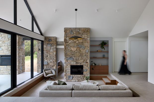 Bright contemporary living room with a large low-profile sectional sofa arranged around a central stone-clad fireplace. Floor-to-ceiling black-framed glazing lines the left side, opening views toward a stone exterior wall and garden. Built-in timber shelving and cabinetry occupy the rear wall, while a sculptural pendant hangs above the fireplace. The room has a high, sloping ceiling and a calm neutral palette with natural stone and timber accents.