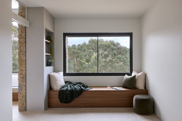 A calm, minimalist built-in window seat occupies the centre of a bright room. The seat has dark timber joinery with integrated storage, neutral cushions, and a deep green throw and cushion. A large black-framed window overlooks trees, while a rough stone structural column and recessed shelving are visible on the left. The room has smooth pale walls, a simple flat ceiling, and light flooring.