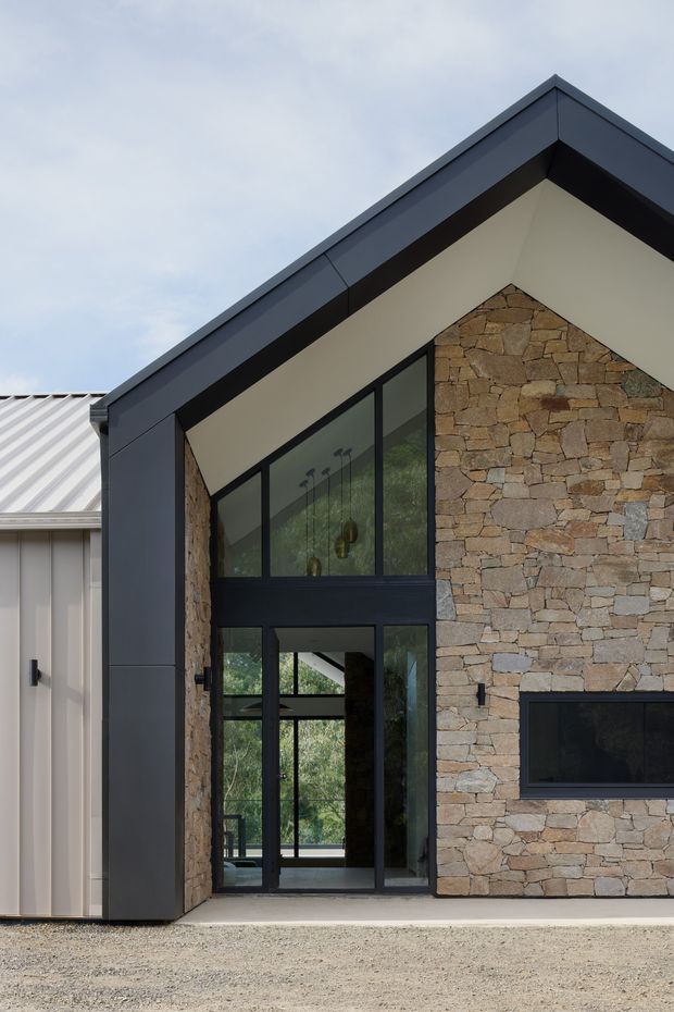 Exterior view of a contemporary gabled house entrance. The facade combines dark metal cladding and window frames with a large irregular natural-stone wall, a tall glazed entry, and a low horizontal fireplace opening. A lighter ribbed metal-roofed wing is visible to the left.
