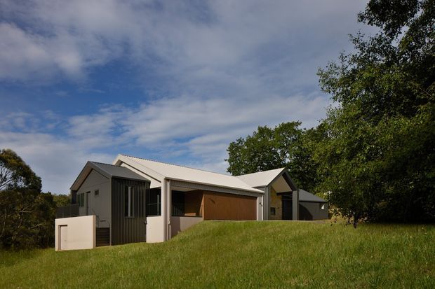 A contemporary single-storey residential building set within a grassy, tree-lined site. The house has intersecting pitched roof forms with light metal roofing, dark vertical wall cladding, pale rendered walls, and a partially recessed covered outdoor area. The image is an exterior architectural view photographed in daylight.