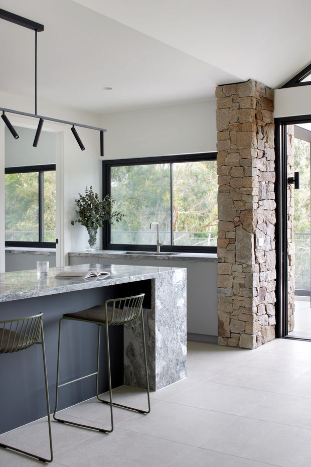 Bright contemporary kitchen interior with a large stone island and integrated breakfast bar, dark cabinetry, expansive black-framed windows, a substantial exposed natural-stone structural column, and a glazed exterior doorway. Minimal black linear pendant lighting and pale flooring create a clean, open feel.